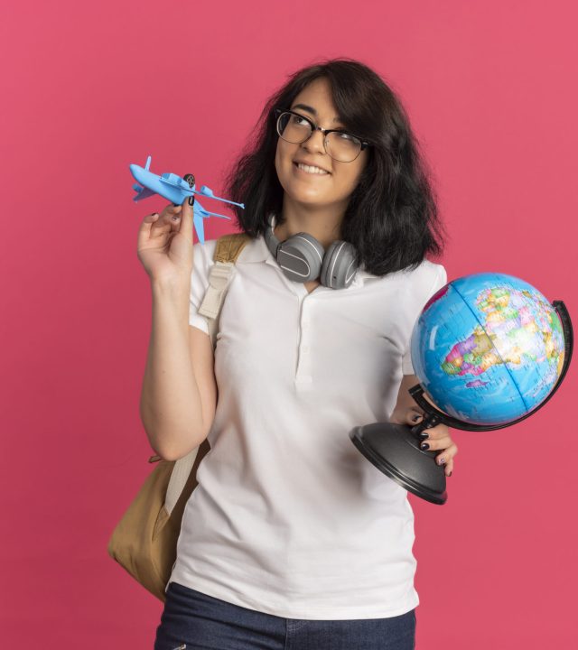 young pleased pretty caucasian schoolgirl with headphones on neck wearing glasses and back bag holds plane and globe looking up isolated on pink background with copy space